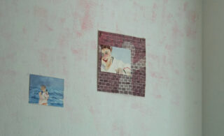 An einer hellen, leicht rosa gefärbten Wand sind zwei Gemälde aufgeklebt; das eine zeigt eine Person vor blauem Hintergrund, das andere eine Person vor einem Backsteinfenster.