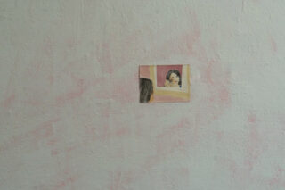 Ein kleines Gemälde einer Person, die ihr eigenes Spiegelbild betrachtet, ist an einer strukturierten, blassrosa Wand angebracht.