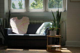 Am Fenster steht ein Sofa mit einer Pflanze und einem Kissen, daneben ein kleiner Beistelltisch mit einer Topfpflanze; Sonnenlicht strömt in den Raum.