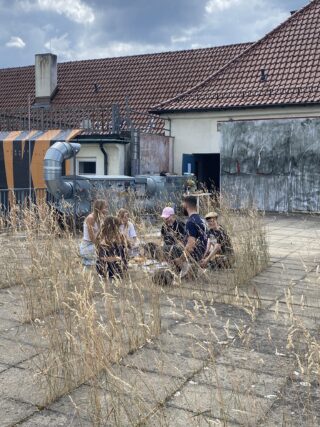 Eine kleine Gruppe von Menschen sitzt zusammen auf dem Boden, umgeben von hohem Gras, auf einem Betondach in der Nähe von Industrierohrleitungen und einem Gebäude mit roten Ziegeln.
