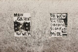 Zwei handgeschriebene Plakate in deutscher Sprache mit schwarzer Farbe sind an einer rauen, grauen Wand angebracht. Auf dem linken Plakat steht 