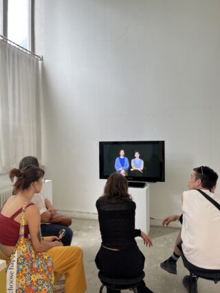 Fünf Personen sitzen auf Hockern vor einem Fernsehbildschirm in einem weiß getünchten Raum und sehen sich ein Video an, das zwei Personen vor einem schwarzen Hintergrund zeigt.