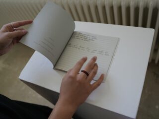 Eine Person mit Ringen an den Fingern blättert in einem kleinen Notizbuch mit handgeschriebenem Text, das auf einem weißen Sockel neben einem Heizkörper liegt.