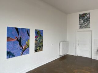 Drei farbenfrohe abstrakte Gemälde sind an einer weißen Wand in einem Raum mit braunem Boden, weißem Heizkörper und weißer Tür ausgestellt.