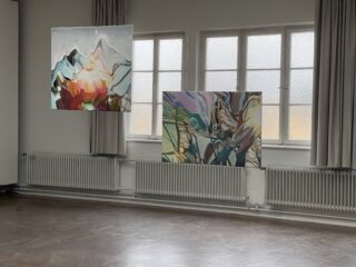 Zwei abstrakte Gemälde sind vor großen Fenstern mit Vorhängen in einem hellen, leeren Raum mit Heizkörpern an der Wand und einem polierten Boden ausgestellt.