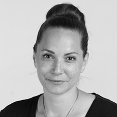 Schwarz-Weiß-Porträt einer Frau mit dunklem Haar in einem Dutt, die ein dunkles Oberteil und eine Halskette trägt und mit neutralem Blick in die Kamera schaut.