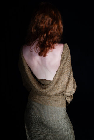 Eine Person mit roten Haaren blickt von der Kamera weg und trägt ein braunes, strukturiertes Oberteil, das ihren Rücken entblößt, vor einem schwarzen Hintergrund.