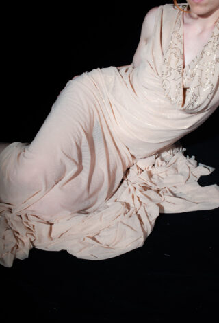 Auf einer schwarzen Fläche liegende Person, die ein langes, drapiertes, ärmelloses beigefarbenes Kleid mit bestickten Details auf dem Mieder trägt.