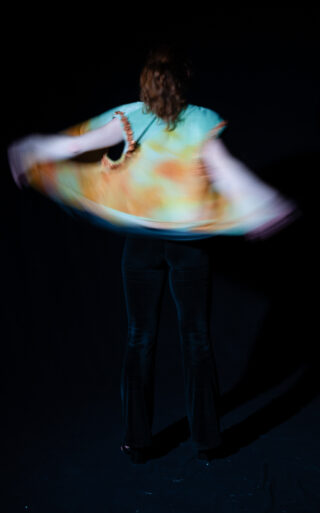 Eine Person mit langen Haaren, von hinten gesehen, dreht sich, während sie ein buntes Oberteil und eine dunkle Hose vor einem dunklen Hintergrund trägt. Durch die Bewegung verschwimmen die Ränder des Oberteils.