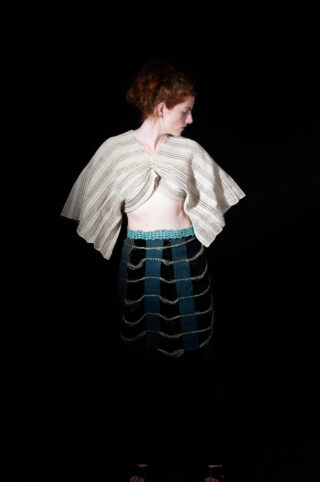 Eine Person mit roten Haaren steht vor einem schwarzen Hintergrund, trägt ein cremefarbenes, strukturiertes Oberteil und einen dunklen Rock mit blauen und goldenen Akzenten und schaut nach links.