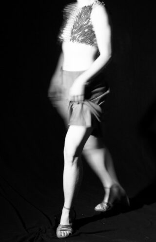 Ein unscharfes Schwarz-Weiß-Foto einer Person in einem strukturierten Oberteil, Rock und Sandalen, die vor einem dunklen Hintergrund geht. Ihr Gesicht und Teile ihres Körpers sind durch Bewegungsunschärfe verdeckt.