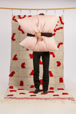 Eine Person steht vor einem strukturierten Teppich mit roten Mustern und hält ein großes rosa Kissen in der Hand, das ihr Gesicht und ihren Oberkörper verdeckt.