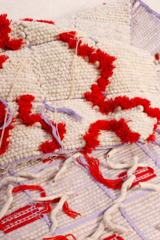 Nahaufnahme eines strukturierten gewebten Textils mit weißem, rotem und lavendelfarbenem Garn, das Tufting- und Knotenmuster und lose Fäden aufweist.