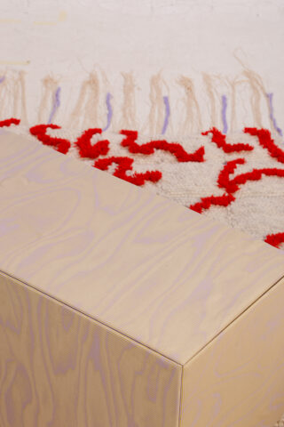 Ein heller rechteckiger Holzblock liegt neben einem cremefarbenen Teppich mit roten und violetten Mustern und Quasten.