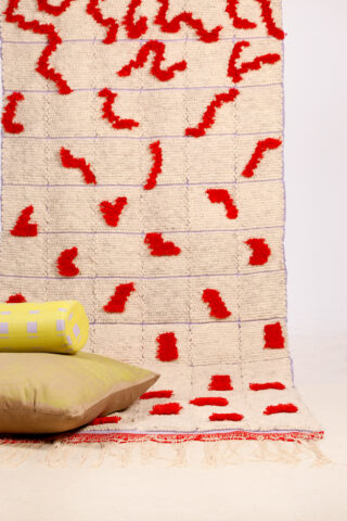 Ein beiger Webteppich mit roten abstrakten Mustern ist vertikal angeordnet, davor liegen ein gelbes zylindrisches Kissen und ein hellbraunes quadratisches Kissen auf dem Boden.