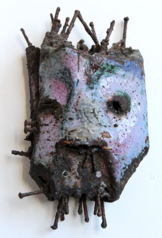 Eine verwitterte, abstrakte Maske mit Pastellfarben und rostigen Nägeln, die aus ihrer Oberfläche herausragen, montiert vor einem weißen Hintergrund.