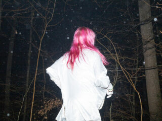 Eine Person mit langem rosa Haar und weißem Hemd steht nachts in einem dunklen Wald und blickt von der Kamera weg, wobei kahle Äste und Schwebeteilchen sichtbar sind.