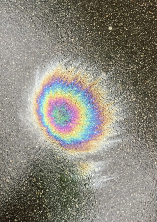 Ein Ölfleck auf nassem Asphalt erzeugt aufgrund von Lichtbrechung und Dünnschichtinterferenz ein kreisförmiges regenbogenfarbenes Muster.