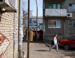Eine Frau geht an einem alten Wohnhaus und einem roten Auto vorbei, an der Wand hängen Plakate und Aushänge, im Hintergrund ist ein Innenhof zu sehen.