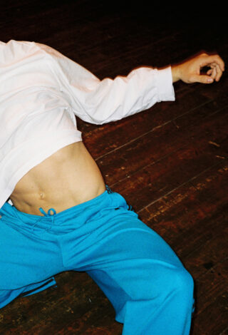 Eine Person, die ein weißes Hemd und eine blaue Hose trägt, liegt auf einem Holzboden, ihr Oberkörper und ein Arm sind im Bild zu sehen.