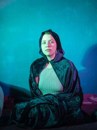 Eine Person, die einen dunklen Samtumhang mit Kapuze trägt, sitzt im Schneidersitz auf einem Bett, das von tealfarbenem und violettem Licht vor einer strukturierten blauen Wand beleuchtet wird.