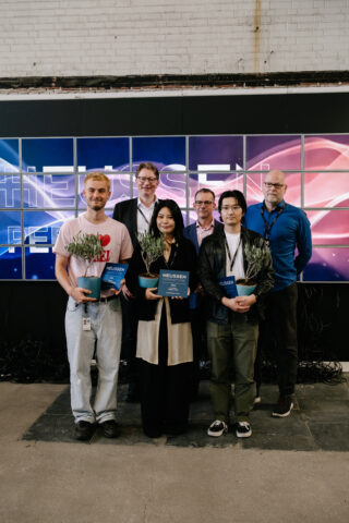 Sechs Personen posieren für ein Gruppenfoto in einem Innenraum vor einer digitalen Anzeige. Drei von ihnen halten Pflanzen und blaue Urkunden.