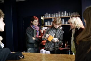 Drei Personen stehen an einem hölzernen Tresen, während eine ein Getränk in einen Becher gießt; im Hintergrund sind Regale mit Gläsern zu sehen.