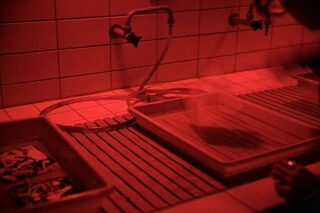 Ein Foto einer Dunkelkammer mit roter Beleuchtung, das Waschschalen auf einem gekachelten Tisch und Filmentwicklungsgeräte zeigt.