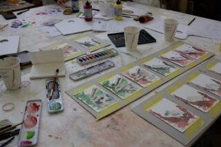 Eine Tischplatte mit Malutensilien, Aquarellpaletten, Pinseln und mehreren kleinen Aquarellbildern, die auf Tafeln aufgeklebt sind; auch Pappbecher und Notizen sind zu sehen.
