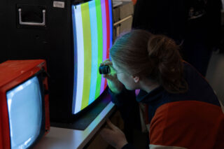 Eine Person untersucht einen Röhrenfernseher, der vertikale Farbbalken anzeigt, mit einer Lupe; ein zweiter Röhrenfernseher zeigt links ein Rauschen an.