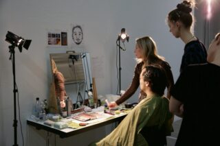 Drei Frauen sind um eine gut beleuchtete Schminkstation mit verschiedenen Kosmetika versammelt, an der eine Frau sitzt, während eine andere das Make-up aufträgt. An der Wand hinter dem Spiegel sind Zeichnungen und Notizen angebracht.