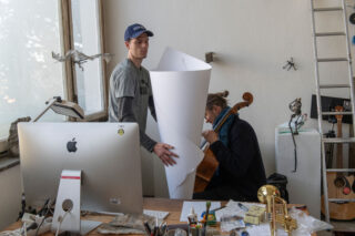 Zwei Personen arbeiten in einem Kunstatelier; eine hält ein großes gerolltes Blatt Papier in der Hand, während die andere mit einem Cello sitzt, das teilweise durch das Papier abgeschirmt wird. Der Schreibtisch ist vollgestopft mit Kunstzubehör und Elektronik.