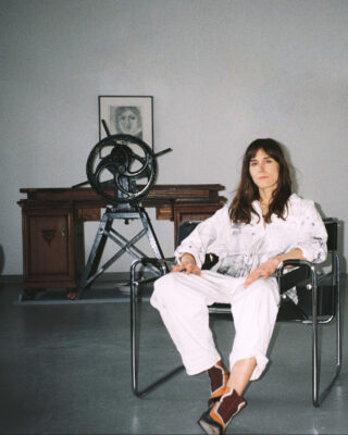 Eine Frau in weißer Kleidung sitzt auf einem modernen Stuhl in einem Raum mit einem Holzschreibtisch, einem großen mechanischen Gerät und einem gerahmten Porträt im Hintergrund.