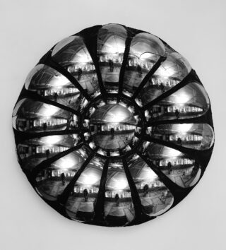 Ein runder, metallischer, mehrteiliger Konvexspiegel, der an einer Wand montiert ist, reflektiert ein verzerrtes Bild eines Innenraums und mehrerer Personen.