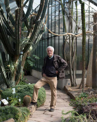 Ein älterer Mann steht auf einem Steinweg in einem Gewächshaus, umgeben von verschiedenen Kakteen und Wüstenpflanzen.