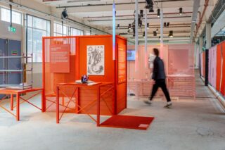 Ausstellungsraum im industriellen Stil mit orangefarbenen Gittertafeln, Tischen mit Büchern und Kunstwerken an der Wand; man geht an den Auslagen vorbei.