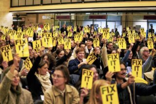 Eine große Gruppe von Menschen, die in einem Raum sitzt, hält während einer öffentlichen Versammlung oder eines Protests gelbe Schilder mit der Aufschrift 