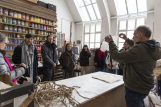 Eine Gruppe von Menschen beobachtet einen Mann, der eine Technik mit weißem Material in einer Werkstatt vorführt, die mit Regalen mit farbigen Gläsern und natürlichem Licht aus großen Fenstern gefüllt ist.