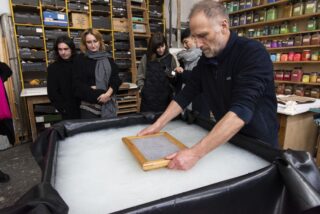 Ein Mann demonstriert die traditionelle Papierherstellung mit einem Holzrahmen, während eine kleine Gruppe von Menschen in einer Werkstatt mit Regalen voller beschrifteter Materialien zusieht.