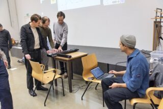 Vier Männer sind um einen Tisch mit einer Tastatur und einem Laptop versammelt. Drei von ihnen stehen und interagieren mit der Tastatur, während einer auf dem Laptop sitzt und tippt. Sie befinden sich in einem geräumigen Innenraum.