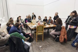 Eine Gruppe von Menschen sitzt und steht um einen großen Tisch mit Speisen und Getränken in einem weißgetünchten Raum und scheint eine Diskussion oder ein Treffen zu haben.