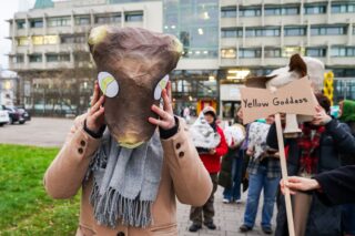 Eine Gruppe von Menschen, einige mit Tiermasken, nimmt an einer Veranstaltung im Freien teil; einer hält ein Schild mit der Aufschrift 