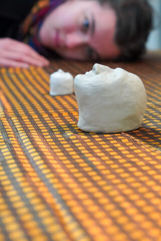 Eine Person liegt mit dem Kopf auf einer gemusterten Fläche und blickt auf zwei kleine Kopfmodelle, die vor ihr aufgestellt sind.