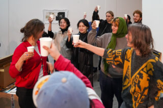 Eine Gruppe von Menschen in Innenräumen stößt mit Pappbechern an, lächelt und unterhält sich in einer ungezwungenen Atmosphäre.