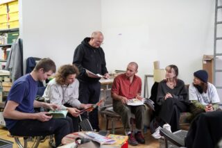 Sechs Personen sitzen und stehen in einem lockeren Workshop-Raum, schauen sich Zeitschriften und Papiere an und diskutieren.