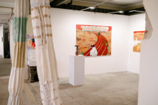 Eine Kunstgalerie mit farbenfrohen Gemälden an weißen Wänden, einer kleinen Skulptur auf einem Sockel und strukturierten Stoffen im Vordergrund.