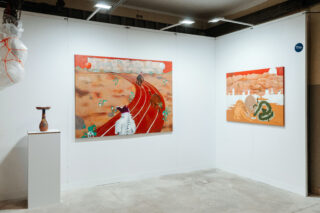 In einer Galerieecke sind zwei farbenfrohe Gemälde mit surrealen Landschaften und eine kleine skulpturale Vase auf einem Sockel zu sehen, alles vor weißen Wänden unter hellem Licht.