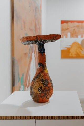 Eine Keramikskulptur mit einer breiten, flachen Oberseite und einem schmalen Sockel, die mit orangefarbenen, gelben und schwarzen abstrakten Mustern bedeckt ist und auf einem weißen Sockel in einer Kunstgalerie steht.