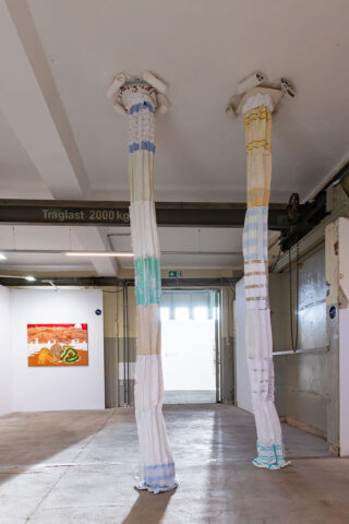 Zwei hohe, stoffbespannte Säulen reichen von der Decke bis zum Boden in einem industriellen Galerieraum; an der Wand im Hintergrund sind Kunstwerke zu sehen.