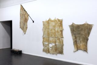 Drei große, unregelmäßig geformte, bräunliche Blätter, die an Tierhäute erinnern, sind an einer weißen Galeriewand angebracht; ein Koffer steht auf dem Boden in der Nähe eines Durchgangs.